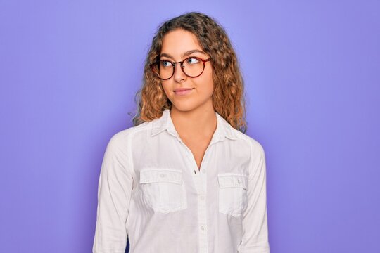 Young beautiful woman with blue eyes wearing casual shirt and glasses over purple background smiling looking to the side and staring away thinking.