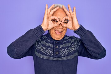 Middle age senior grey-haired man wearing glasses and winter sweater over purple background doing ok gesture like binoculars sticking tongue out, eyes looking through fingers. Crazy expression.