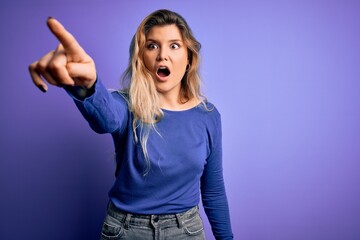 Fototapeta premium Young beautiful blonde woman wearing casual t-shirt over isolated purple background Pointing with finger surprised ahead, open mouth amazed expression, something on the front