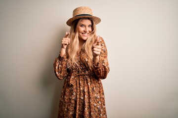 Young beautiful blonde woman wearing summer dress and hat over isolated white background pointing...