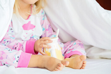 Obraz na płótnie Canvas Cute little girl with blonde hair sitting on the bed and drinking milk. Happy childhood. Stay at home during coronavirus covid-19 pandemic quarantine concept.