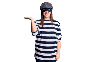 Young beautiful brunette woman wearing burglar mask smiling cheerful presenting and pointing with palm of hand looking at the camera.