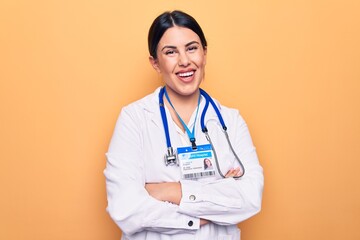 Young beautiful doctor woman wearing stethoscope and id card over isolated yellow background happy face smiling with crossed arms looking at the camera. Positive person.