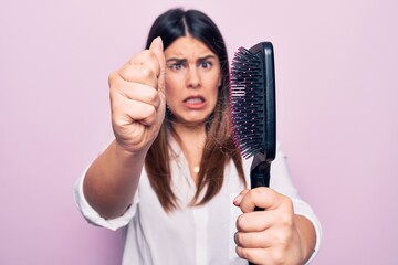 Young beautiful woman agry and worried for capillary problem. Holding hairbrush with tangled hair standing over isolated pink background