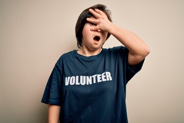 Young down syndrome volunteer woman wearing social care charity t-shirt peeking in shock covering face and eyes with hand, looking through fingers with embarrassed expression.