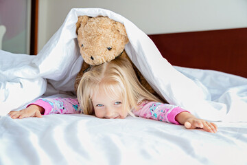 Cute little girl with blonde hair lying on the bed with stuffed teddy bear. Happy childhood. Stay at home during coronavirus covid-19 pandemic quarantine concept.