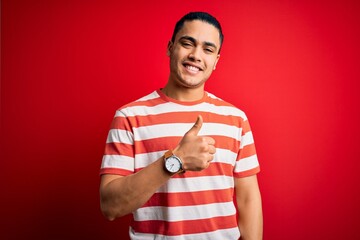 Young brazilian man wearing casual striped t-shirt standing over isolated red background doing happy thumbs up gesture with hand. Approving expression looking at the camera showing success.