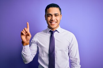 Young brazilian businessman wearing elegant tie standing over isolated purple background showing and pointing up with finger number one while smiling confident and happy.
