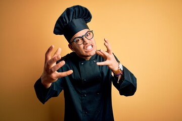 Young brazilian chef man wearing cooker uniform and hat over isolated yellow background Shouting frustrated with rage, hands trying to strangle, yelling mad