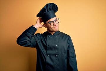 Young brazilian chef man wearing cooker uniform and hat over isolated yellow background confuse and wondering about question. Uncertain with doubt, thinking with hand on head. Pensive concept.