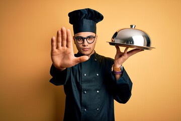 Young brazilian chef man wearing cooker uniform and hat holding tray with dome with open hand doing stop sign with serious and confident expression, defense gesture
