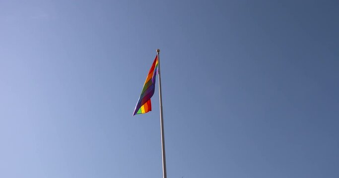 Rainbow Pride flag gently swaying against a blue sky background.
