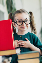 Smiling little blonde kid girl in round shaped glasses 4-6 years old in library among many books. Kid education concept