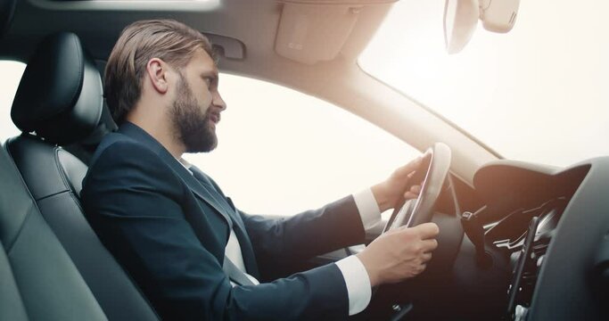 Busy bearded businessman in formal outfit driving car and using loudspeaker on smartphone for conversation. Mature man enjoying modern technology to have free hands in auto.