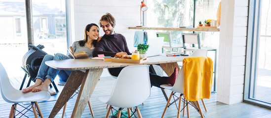 Happy couple working together at home. Sitting at the table with laptop and snacks. Reading book and using computer. Wide screen, panoramic
