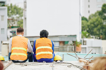 Construction workers in bright orange vests resting after shift and looking at site, view from the...