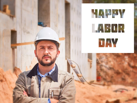 Happy Labor Day. Caucasian bearded civil engineer or construction worker in white hardhat stands cross handed opposite construction site - Powered by Adobe