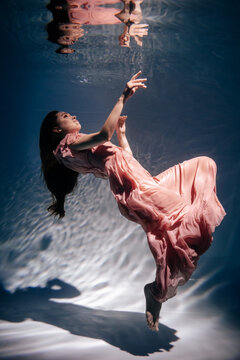 Woman Wearing Fashion Pink Dress Underwater Pool. Beautiful Young Lady Model with Long Hair and Wear Style Clothes Swim in Clean Water. Atmospheric Fairy Tale or Magic Vertical Photo
