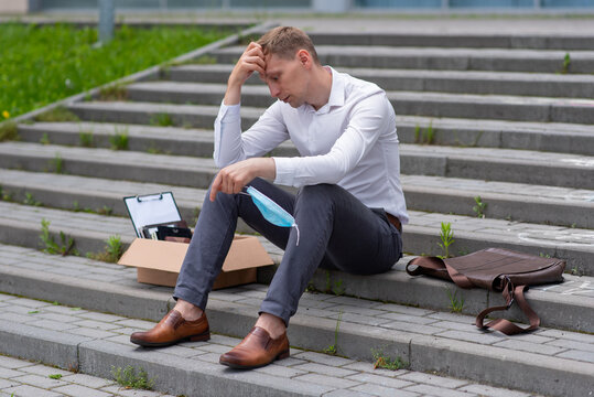 Dismissal Of An Employee Due To A Coronavirus Epidemic. A Man Sits On The Steps. Next To Him Is His Stationery. He Has A Medical Mask In His Hand.