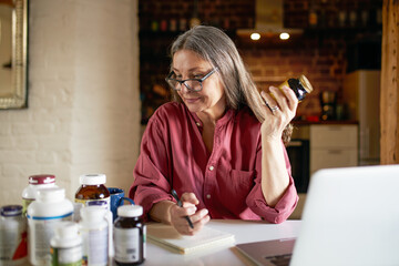 Portrait of middle aged female nutritionist in eyeglasses making research, sitting in front of open...