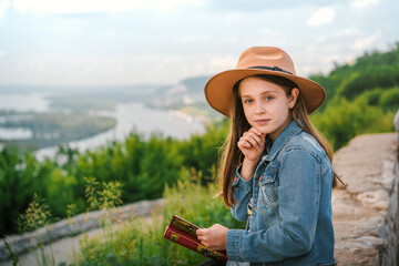 Little child girl sitting in a denim jacket and hat on a rock with an amazing view of the mountains and the Volga river and read the book,  the concept of romance and travel, nature of Russia
