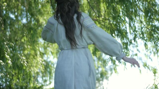 A girl dressed in an old dress stands on the river bank. Beautiful woman standing near a willow tree. Young girl walks in nature alone. Summer nature.