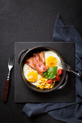 Fried egg with slices of bacon, tomato, corn and spinach black frying pan on a black background....