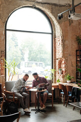 Young male friends resting in loft cafe and using modern devices while discussing last apps