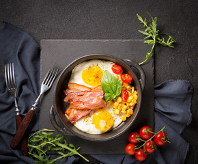 Fried egg with slices of bacon, tomato, corn and spinach black frying pan on a black background. Breakfast. Top view