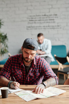 Serious Brutal Guy In Ball Cap Sitting At Table In Cafe And Planning Route With Paper Map