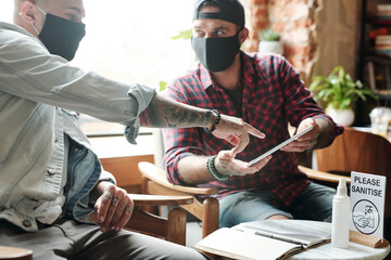 Young men in cloth masks sitting in lobby and discussing new app on tablet