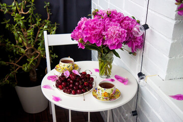 Bouquet of peony flowers, two cups of tea and fresh cherries on white table. Modern balcony interior.