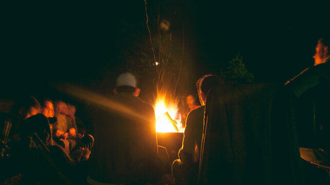 The Group Of Young People Are Sitting Around The Bonfire And Talking And Singing Songs