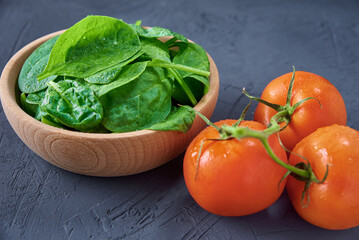 Fresh spinach leaves in wooden bowl and tomato on dark background. Organic food