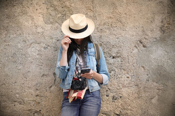 Woman wearing blue jeans and hat, standing against wall and using smartphone