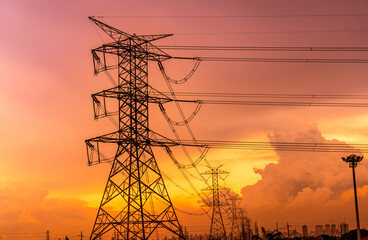 High voltage electric pylons and transmission lines with orange sunset sky and cityscape modern...