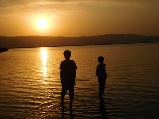 couple of kids at sunset beach standing in water with immersed feet and legs making beautiful silhouette of them. Darkness and soe golden and  lights on sunset horizon of  sky.