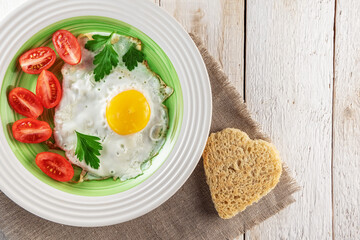 Heart shaped fried egg, tomatoes and bread on white wooden background