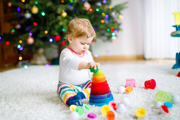 Fototapeta na wymiar Adorable cute beautiful little baby girl playing with educational toys at home or nursery. Happy healthy child having fun with colorful wooden rainboy toy pyramid. Kid learning different skills.