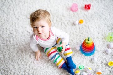 Adorable cute beautiful little baby girl playing with educational toys at home or nursery. Happy healthy child having fun with colorful wooden rainboy toy pyramid. Kid learning different skills.
