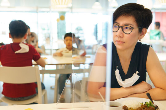 Stylish Middle Aged Asian Woman With Medical Face Mask Sit Separate From Her Kids In Food Court With Clear Acrylic Divider / Barrier On Table. New Normal & Social Distancing During Covid-19 Pandemic