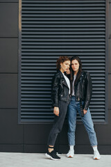 Vertical portrait of two girlfriends in casual clothes, wearing leather jackets and jeans, standing on a black background, posing at the camera.Fashionable couple of girls posing on black wall