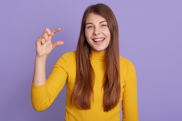 Indoor shot of glad young woman with long beautiful hair, shows very tiny size with fingers, dresses casual clothes, poses isolated over lilac background.