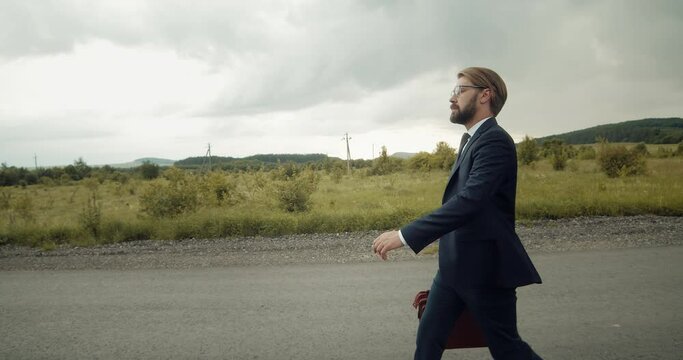 Side view of confident businessman walking on road among green beautiful nature. Bearded man wearing eyeglasses and formal clothing. Successful person carrying on leather bag with important documents.
