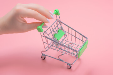 Small shopping cart in female hands. a woman's hand carries a small grocery cart on a pink background. Shopping and retail concept