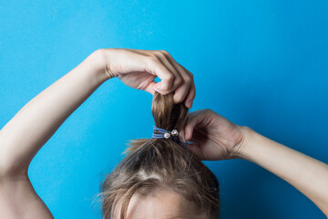 Girl straightens the disheveled tail of hair on her head, against a blue background. Modern fast hairstyle. Hair tied with elastic band