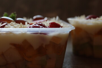 fresh fruit salad of melon, grape, strawberry, kiwi dressed with greek yoghurt and sprinkled with chopped Apple. gratted cheese and fruits for upper topping. served in a square plastic on wooden tray.