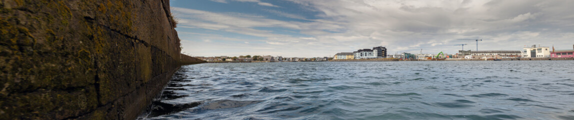 View on Galway city from river Corrib, low angle, panorama image. The long walk and dock area, Warm sunny day, Cloudy sky over city
