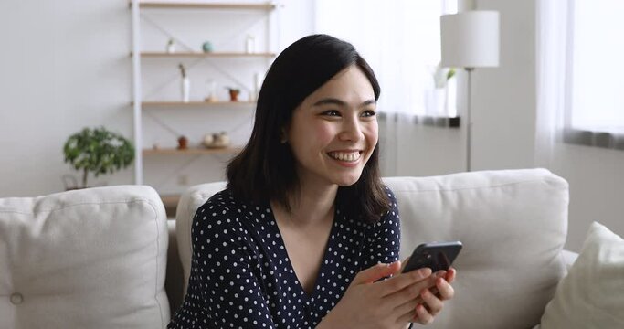 Happy Asian young woman sit on sofa hold cellphone receive message feels excited celebrating on-line lottery victory, got opportunity hired job of dream, new app software, moment of win candid emotion