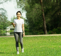 Healthy young beautiful asian woman walking and listening to music on earphone after running or jogging in the park in sunshine on beautiful summer day. Sport fitness training outdoor concept Healthy